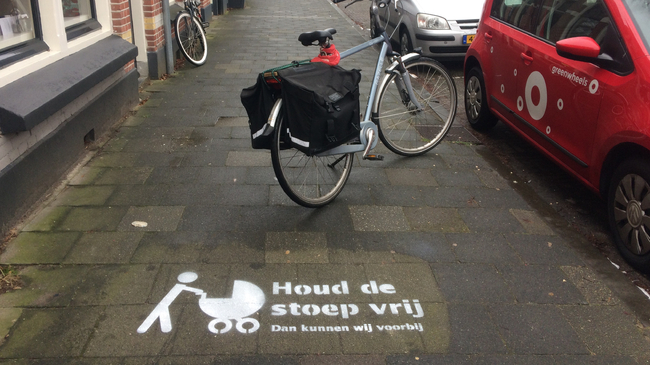 Stope met opschrift 'Houd de stoep vrij' en plaatje van kinderwagen. En een fiets die midden op de stoep is geparkeerd.